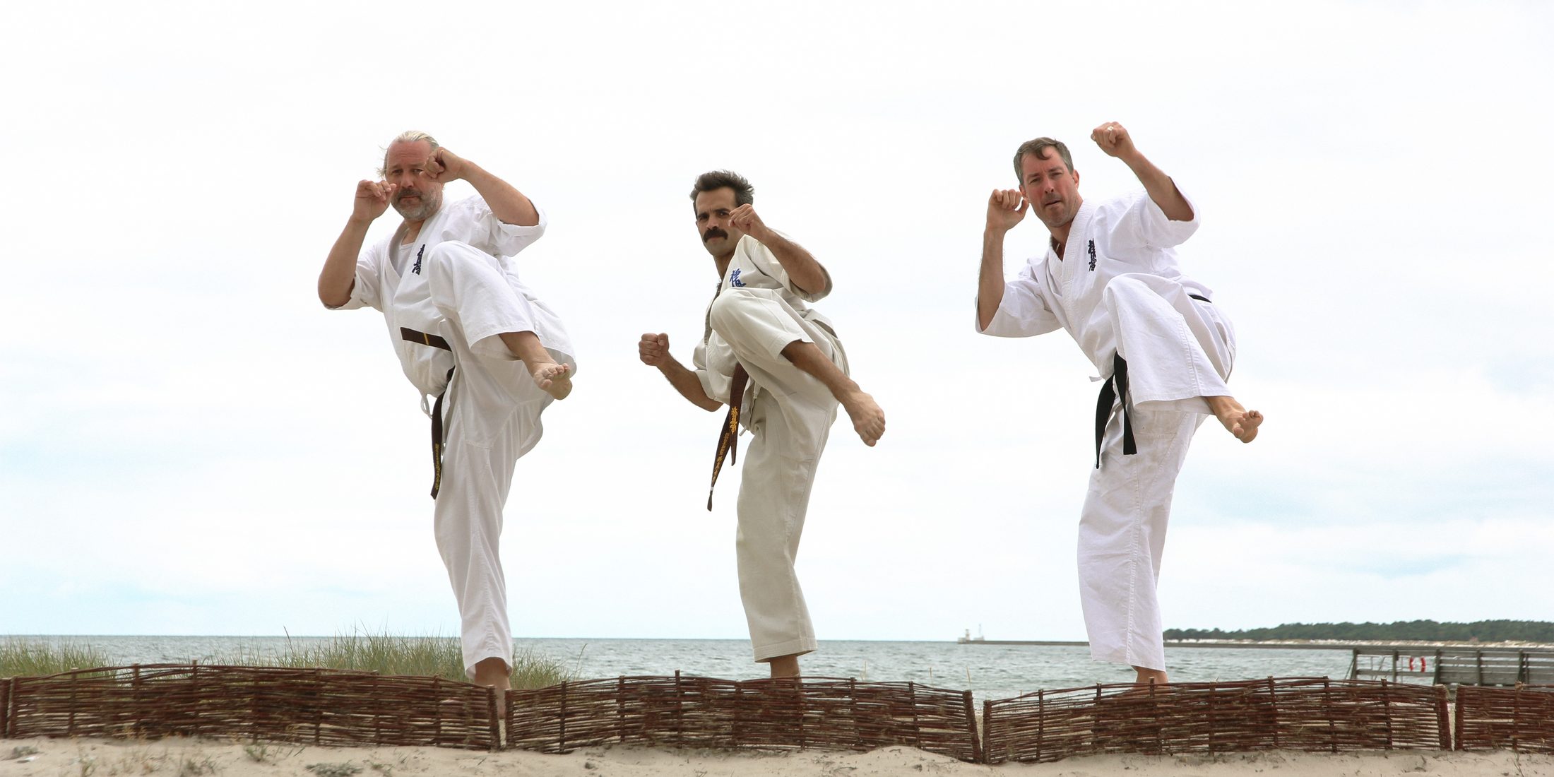 Tre karateka i gi som sparkar högt på stranden med havet bakom sig